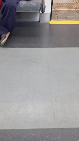 The video shows a person inside what appears to be a public transportation vehicle, possibly a train, with a floor that is being mopped. The floor is gray with speckles. The person is wearing black pants and black athletic shoes. They are holding a mop with a blue and white mop head attached to a light blue pole, and are actively cleaning the floor. In the background, another person is seated on a bench, wearing dark blue pants and brown shoes. The interior of the vehicle shows a gray bench seat and a yellow strip along the edge of a doorway. The scene appears to be indoors. The lighting suggests it could be daytime, but there are no clear indications of the time of day or weather. The activity is routine cleaning. No text is visible in the video.