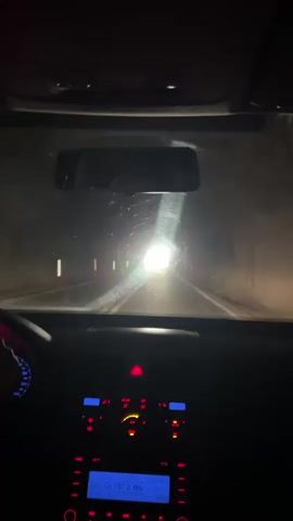 The video shows the interior of a car as it drives through a tunnel. The driver is visible in the rearview mirror. The dashboard and steering wheel are visible in the foreground. The car is moving towards a bright light at the end of the tunnel. The car's radio is visible on the dashboard. The windshield is a bit dirty. A red hazard light is blinking on the dashboard.