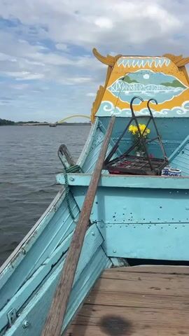 The video opens with a close-up shot of the bow of a light blue wooden boat. A wooden oar leans against the side of the boat. In the background, a calm body of water stretches out towards a distant yellow bridge under a cloudy sky. The boat is decorated with traditional yellow and blue painted designs on its ornate, curved roof. In the center of the bow, a small altar-like structure holds a bunch of yellow flowers and what appears to be incense. As the video progresses, the camera pans slightly to reveal a young boy with brown hair sitting on the wooden deck of the boat. He is wearing a light beige sleeveless shirt. He looks towards the camera and speaks, but his words are not clearly discernible. The boat appears to be moving gently on the water, and the overall atmosphere is peaceful.