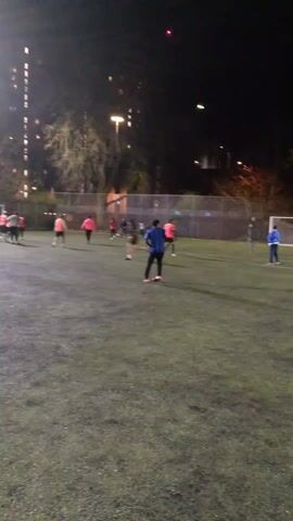 The video shows a group of people playing soccer on a dark, outdoor field. The field has artificial turf and is lit by bright overhead lights. Players are wearing a variety of clothing, including brightly colored vests and jackets. The players are running around the field and kicking a soccer ball. There is a soccer goal visible at one end of the field, and a building and trees can be seen in the background.