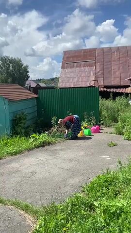 A woman in a dark blue and burgundy outfit is kneeling in the dirt near a green metal fence, appearing to be gardening. She is facing away from the camera and has a yellow watering can and a pink watering can next to her. Behind her is a long, rustic-looking building with a corrugated metal roof. The scene is set outdoors on a sunny day with white clouds in a blue sky.
