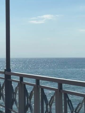 The media file depicts a tranquil scene of a bright, sunny day over a body of water, likely a lake or sea. In the foreground, a white metal railing with an ornate, geometric pattern runs horizontally across the frame, suggesting a pier, boardwalk, or elevated viewing platform. A thick, gray pole, possibly a lamppost or structural support, stands vertically on the left side, partially obscuring the view.

The water's surface is a deep blue and reflects the sunlight, creating a shimmering, sparkling effect. The horizon is visible in the distance, a faint line separating the water from a pale blue sky. A few wispy white clouds are scattered across the upper portion of the sky. The overall impression is one of peacefulness and natural beauty.

The time of day appears to be late morning or early afternoon, given the bright sunlight and clear sky. The weather is fair and sunny. The location context provided is Koshkol', Kyrgyzstan, which is known for its location on the southern shore of Issyk-Kul Lake, a large alpine lake that matches the visual cues in the media. There is no visible text in the media file. The scene evokes a sense of calm and relaxation, inviting contemplation of the vastness of the water and sky.