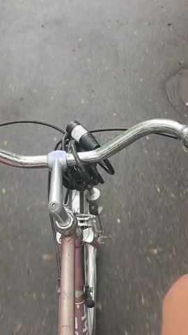 The video shows a first-person view from the handlebars of a bicycle moving on asphalt. A hand appears holding a clear plastic container with several pieces of dessert that look like baklava. The person states in Bulgarian that they are burning calories and that there is no chance. The container is then placed back on the handlebars.