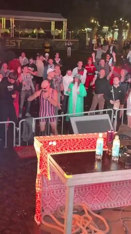 The video shows a crowd of people at an outdoor event at night. A man in a pink and black striped singlet and shorts is standing in front of a metal barrier, facing the crowd and holding his fists up as if in a boxing stance. He appears to be bald and is wearing sunglasses. Behind him, other people are watching, some with their hands raised. In the foreground, a table is covered with a red and gold patterned cloth, with several bottles and two white objects on it. The environment appears to be festive, with some lighting and structures visible in the background. The overall mood seems to be energetic and participatory.