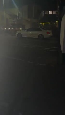 A white sedan is parked in a dimly lit outdoor space. The car is positioned at an angle to the viewer. The background shows buildings with lights on, suggesting it is nighttime. There are some trees or bushes visible in the background. The ground appears to be paved, possibly a parking lot, with painted lines. The overall lighting is low, with some areas brighter due to artificial lights.