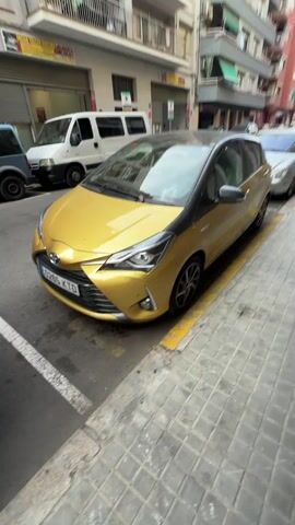 A golden Toyota Yaris is parked on a street next to a sidewalk. The car is facing the camera and is positioned in a parking spot marked with white lines. The car has a black roof and black trim around the windows. The car's headlights are on. In the background, there is a white van and a blue van. There are also buildings with windows and some signage. The street is paved and there is a yellow line along the edge of the sidewalk. The video appears to be shot from a moving vehicle.