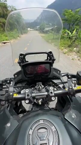 The video shows a first-person perspective from a motorcycle rider, interspersed with a view of the rider and their passenger.

The initial shot is from the rider's perspective, looking forward down a two-lane asphalt road with a solid yellow center line. Lush green, forested hillsides flank the road, which appears to curve gently. In the distance, several simple houses or buildings are visible amidst the mountains. The motorcycle's windshield, dashboard (displaying "50" briefly), handlebars, and mirrors are prominent. The handlebar clamps have "PANTARACING" written on them.

The camera then shifts to a selfie-style shot, showing the rider and a passenger from the front. The rider wears a red jacket, sunglasses, and a white and yellow helmet with a DJI Osmo Action camera mounted on the chin. The passenger, seated behind the rider, wears a black t-shirt, sunglasses, and a gray helmet, with one arm raised high, holding a smartphone as if taking a picture or video. They both appear to be enjoying the ride.

The final shot returns to the first-person perspective, continuing along the road. The sky is partly cloudy, and the overall impression is of a scenic ride through a mountainous, rural area. A white road sign with some Vietnamese text ("ĐIỂM TR") is briefly visible on the left side of the road at the 0:02 mark.