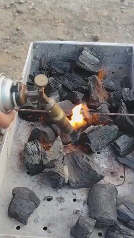 The video shows a close-up, overhead view of a person's hands using a propane torch to ignite charcoal briquettes in a rectangular metal grill. The grill contains a substantial amount of black charcoal, some of which is already glowing red and producing smoke. The torch flame is directed at a pile of charcoal, causing it to ignite and flare up. The background is blurred and appears to be an outdoor, possibly dusty environment. The person's hands are positioned to hold the torch and manipulate it to ensure the flames spread. The action is focused on the process of starting a charcoal fire.