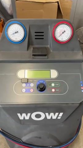 The video shows a close-up of a WOW! air conditioning refrigerant machine. The machine is gray and has two pressure gauges on top, one with a blue rim and the other with a red rim. There is a small display screen in the center of the machine, with buttons and a knob labeled "ENTER" below it. The words "WOW!" are printed in large white letters on the bottom of the machine. A hand enters the frame and points to the word "WOW!" on the machine. The hand then moves out of the frame. The background shows boxes and other equipment, suggesting the setting is a garage or workshop.