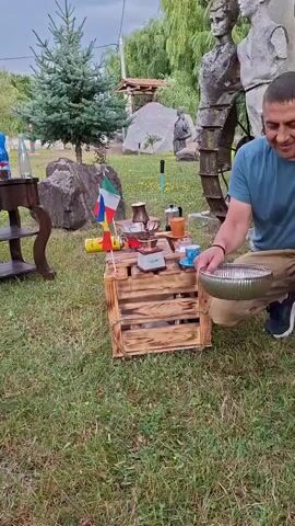 A man is on his knees in a grassy, outdoor area. He is wearing a blue t-shirt and tan pants. He is holding a silver bowl in his right hand and appears to be interacting with a small, wooden crate in front of him. The crate is adorned with a small Italian flag and is holding a coffee-making apparatus, including a metal pot and a small cup. Other objects in the background include a shelf with bottles, large rocks, a evergreen tree, and several abstract sculptures.