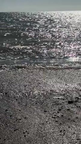 The video presents a static, ground-level view of a beach where gentle waves are lapping against the shore. The water glitters with sunlight, creating a shimmering effect across the surface. The beach appears to consist of dark sand and small pebbles. The horizon line is visible in the distance, with a pale sky above.