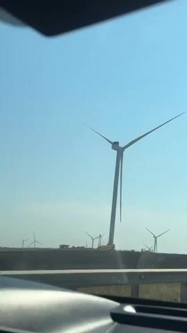 The video shows a view from a vehicle traveling on a road. In the foreground, a guardrail is visible. In the background, several wind turbines are present, some appearing to be under construction. The sky is clear with some scattered clouds, and the sun is shining brightly. The landscape appears to be flat with some sparse vegetation. A traffic sign indicating a sharp curve to the left is also visible. The vehicle is moving forward, and the scenery is captured from a moving perspective.