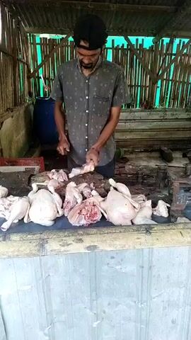 A man in a dark patterned shirt and a black cap stands behind a counter, wearing a dark apron. He holds a knife in his right hand and appears to be butchering raw chickens on a cutting board. Several whole and partially cut chickens are displayed on the counter in front of him. The scene appears to be an outdoor market stall with a corrugated metal roof and bamboo walls. A large blue barrel and a red plastic crate are visible in the background. The overall impression is of a food preparation area.