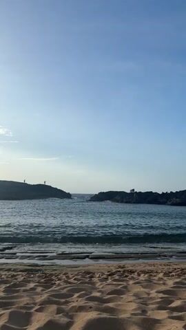 The video shows a view from a sandy beach towards the ocean. The sky is clear and blue, with the sun low in the sky casting a soft light. In the distance, two rocky outcroppings are visible, separated by a channel of water. Several people are standing on the rocks, and there is a flag flying on the right outcropping. Gentle waves are breaking on the shore in the foreground. The overall atmosphere is serene and peaceful.