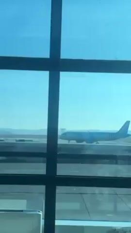 The video shows an airplane on a runway viewed through a window with a grid-like frame. The plane is light blue and has the words "FLYONE" on the tail and body. It appears to be stationary. The sky is clear and blue. The runway is grey, and there are other planes in the distance. The view is slightly obscured by the window frame.