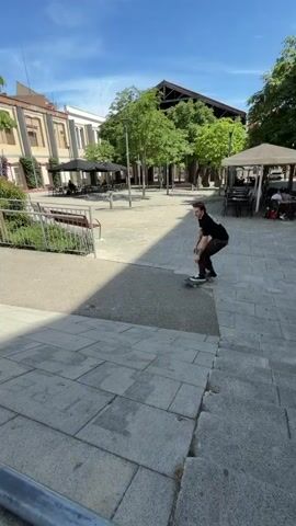 A skateboarder with tattoos on his arms, wearing a black t-shirt and brown pants, attempts a trick on a metal rail. The skateboarder rides down a paved slope, with buildings and trees in the background. The skateboarder lands on the rail and slides, but then falls off, and the skateboard rolls away. The scene appears to be an outdoor urban area.