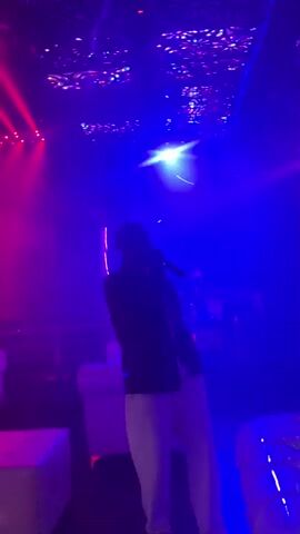 A Black male wearing a dark jacket and light-colored pants stands in the center of a dimly lit room, holding a microphone. He is facing forward and appears to be singing or speaking. The room is decorated with colorful lights on the ceiling, which project patterns onto the walls. The overall atmosphere suggests a nightclub or concert venue.