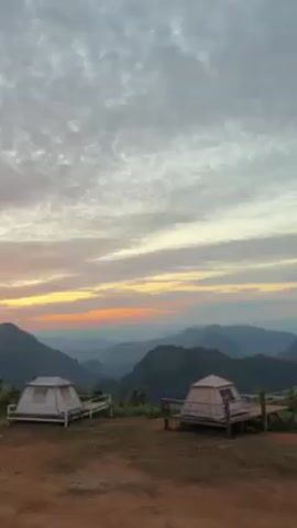 The video shows two white tents on wooden platforms on a hilltop, with a view of mountains in the distance. The sky is cloudy with a pink and orange sunset. People come into the frame in front of the tents. There is a conversation occurring.