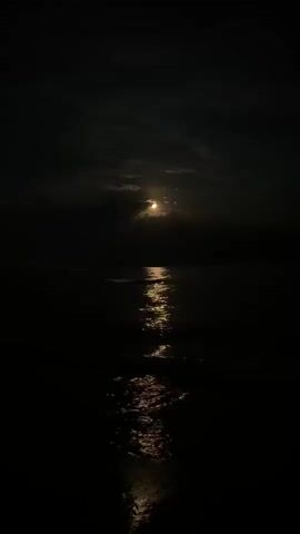 The video shows a dark, nighttime scene over a body of water, likely the ocean. A full moon is visible in the upper center of the frame, partially obscured by clouds. The moonlight reflects brightly on the surface of the water, creating a shimmering path that extends towards the viewer. Gentle waves are visible, contributing to the reflective pattern. The overall impression is one of peaceful, natural beauty.