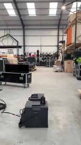 A black fog machine emits a plume of white fog into the air in a large warehouse-like space. The machine is in the foreground, connected to a power cord. Behind it, various pieces of equipment are visible, including black cases on wheels, cables, and storage racks with boxes and other items. The space has a high ceiling with multiple skylights, providing ample light. The floor is a smooth gray concrete.