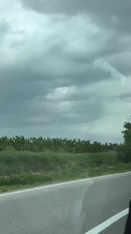 The view from a moving vehicle shows a cloudy sky and roadside vegetation. The road has a white line marking the edge. A utility pole is visible in the distance.
