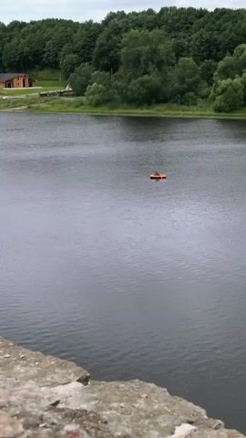 A person in a red shirt is in an orange inflatable boat on a dark body of water. The boat is moving away from the camera. Behind the water is a dense, green forest under a cloudy sky. In the foreground, the edge of a stone structure is visible.