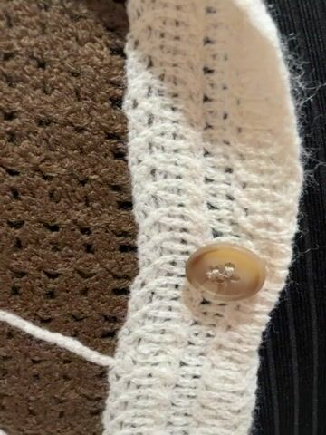 This media file shows a close-up and then a wider view of a brown and white knitted button-up shirt with a collar, displayed on a mannequin. The shirt has a repeating leaf-like pattern in white against the brown knit. The button placket is white and features beige buttons. A black undergarment is visible beneath the open shirt. The wider shots reveal the shirt is on a mannequin in what appears to be a retail store environment, with racks of clothing visible in the background. No people are visible, and the time of day and weather are not discernible. There is no visible text in the media. The scene is set within a retail store, likely a clothing boutique, and given the context provided, it is located in Ko Samui District, Thailand.