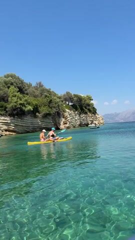 Two people are kayaking in a clear, turquoise ocean. They are paddling a yellow kayak and appear to be enjoying a sunny day. The water is so clear that the rocky seabed is visible. A lush, green cliffside covered in trees rises from the water to their left. In the distance, a boat is anchored and mountains can be seen. The sky is a bright, clear blue.