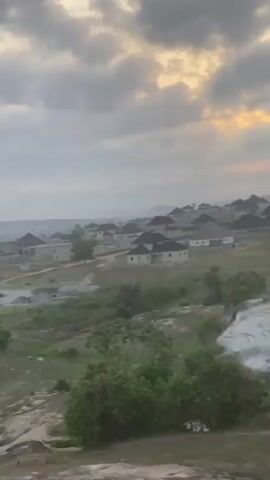 The video shows a landscape on a cloudy day. In the foreground is a rocky terrain with sparse vegetation. In the midground, there is a mix of green fields and some areas that appear to be under construction or recently developed. The houses with dark roofs can be seen in the distance. The sky is overcast with varying shades of gray clouds.