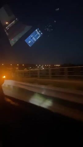 The video shows a view from inside a vehicle at night. The dashboard of the vehicle is visible, displaying a digital screen with various icons and information. Outside the vehicle, a highway is visible with lights from street lamps and possibly buildings in the distance. There is a railing along the side of the highway. The road ahead has white lines marking the lanes. The sky is dark, indicating it is nighttime. The motion of the vehicle is evident from the blur of the lights and the environment outside.