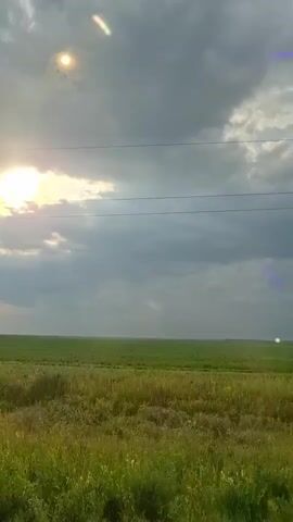 The video shows an outdoor scene with a vast expanse of green fields under a cloudy sky. The sun is visible in the upper left corner, partially obscured by clouds, and appears to be shining brightly. There are two horizontal lines, likely power lines, stretched across the sky. In the middle of the frame, a vertical streak of light, displaying a spectrum of colors from red at the bottom to green at the top, is visible in the distance. There is also a small, bright white circular light in the far right of the frame. The foreground consists of tall, green grass with some smaller plants and flowers. The overall impression is a landscape view with some atmospheric optical phenomena.