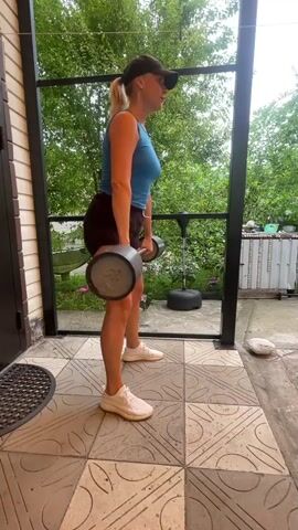 A woman with blonde hair tied in a ponytail, wearing a blue tank top, dark shorts, and a black baseball cap, performs deadlifts with a barbell outdoors on a tiled patio. She is positioned in profile, facing left. In the first frame, she is standing upright with the barbell held in front of her thighs. In the subsequent frames, she bends at the hips and knees, lowering the barbell towards the ground while maintaining a straight back and extended arms. She then returns to the starting standing position. The background shows greenery, a fence, and a punching bag.