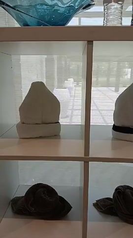 A white shelving unit with four compartments is shown, each compartment containing folded towels. The top row of compartments shows two towels folded into a pointed shape, resembling hats. The towel on the left is solid white, while the towel on the right is white with a black band around the base. The bottom row of compartments shows two towels folded into a spiral shape, resembling snails or roses. The towels in the bottom row are a dark green or brown color. Above the shelving unit, a blue glass bowl and two clear water bottles are visible on a shelf. The background appears to be an interior space with sunlight filtering through blinds, creating a grid pattern on the floor.