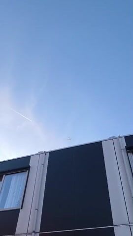 A low-angle, vertical video shows the upper section of a building against a clear blue sky. The building has a flat roofline and appears to be constructed with panels. The wall section visible is divided vertically into three sections: a white panel on the left, a dark gray or black panel in the center, and a white panel on the right. A window with vertical blinds is visible on the left white panel. White pipes run vertically along the seam between the left and center panels. A faint white contrail stretches across the sky diagonally from the lower left to the upper right. The sun is low in the sky, creating a slight lens flare effect on the left side of the frame. The overall impression is a clear, bright day.