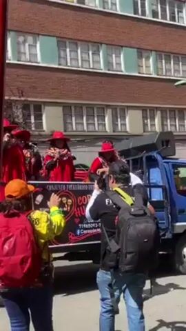 The video captures a lively outdoor scene featuring a band of musicians performing on the back of a blue truck. The musicians are dressed in vibrant red outfits with matching hats. A banner on the truck identifies them as "Conjunto Folklórico Corazon Humano". People around the truck are recording and enjoying the performance. The backdrop features a brick building with numerous windows. A red pole is visible on the left side of the frame, partially obscuring the view. The weather appears sunny, creating a bright ambiance.