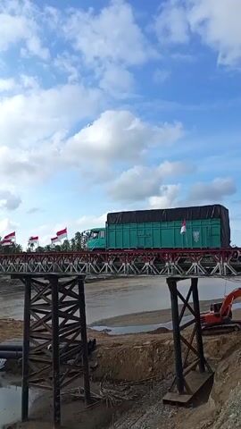 The video shows a teal cargo truck driving over a truss bridge, decorated with Indonesian flags, against a partially cloudy, blue sky. The bridge appears to be constructed of red and silver metal, with wooden supports. An excavator sits idle on the dry ground beneath the bridge, next to some black pipes, in what looks like a construction zone. Some puddles of water lie between the bridge’s supports.