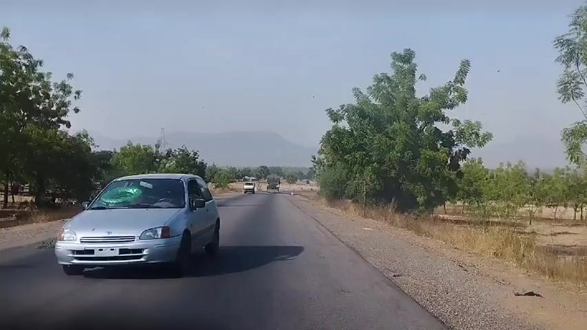 The video depicts a scene of a car driving on an asphalt road during the day. The road is flanked on both sides by trees and dry, grassy terrain. In the distance, mountains are visible under a clear sky. Other vehicles, including a white van and a larger truck, can be seen further down the road. A motorcycle is visible on the left side of the road.