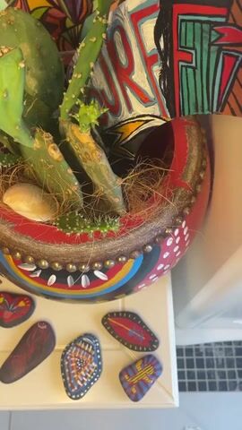 The video opens with a close-up of a decorative planter containing a cactus and surrounded by painted stones. The planter is brightly colored with a red rim and white dots. The stones, also vibrantly painted with various designs, are scattered on a white surface. The camera then pans to the right, revealing a white dresser. On top of the dresser, there is a vintage-style fan and a gold circular tray adorned with a painted bird and colorful rays. The tray sits on a lime-green, textured surface, which holds several watches and rings. More painted stones are visible on the white surface below. The video continues to pan across the dresser, showing glimpses of other objects including a green bottle opener and a blurred background of what appears to be a shelf with various items. The final shot focuses on the gold tray and the jewelry on the green surface.