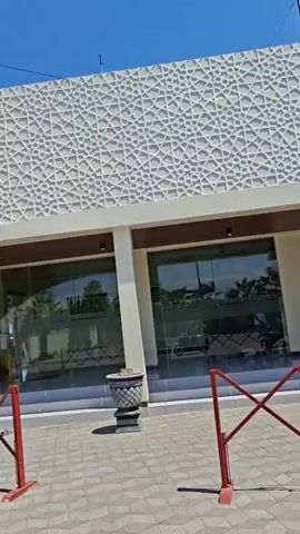 The video shows the exterior of a building with the words "MASJID BILFAQIH ZAIN" displayed prominently. The building features modern architecture with large glass windows and a distinctive decorative facade composed of a repeating geometric pattern. In the foreground, a red collapsible barrier is visible, partially obstructing the view of the entrance. A paved walkway leads to the building, and a large grey urn is positioned to the right of the entrance. The sky is clear and blue, suggesting fair weather. There are trees and some utility poles in the background, and a gray car is seen driving away from the building. Some people are seated on steps near the entrance, and a yellow information kiosk with a QR code is also present. The overall impression is of a contemporary public building, possibly a mosque, under a bright sky.