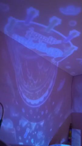 A purple light with various light blue patterns, including the words "Happy Birthday" and a cake, is projected onto a white wall and ceiling in a room.