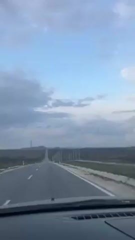 The video shows a car driving down a straight, empty road under a partly cloudy sky. There's a line of tall, white posts on the right side of the road, and flat, grassy land on both sides. A power line tower is visible in the distance. The interior of the car is also visible, with the dashboard in the foreground. The video has accompanying music.