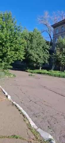 The video shows an outdoor scene with buildings and trees under a clear blue sky. The camera pans across a dusty, cracked asphalt area. In the first frame, a multi-story brick building is visible on the right, with trees in front of it. As the camera moves, more of the building and surrounding greenery are revealed. In the later frames, the sun is bright, casting light and creating lens flares. Other buildings and trees are visible, and the ground continues to show signs of wear and tear. There are no people clearly visible in the footage.