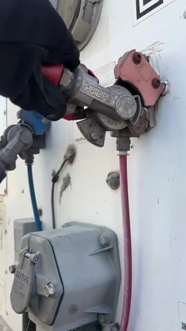 The media file captures a close-up, low-angle shot of a gloved hand connecting a red air hose to a trailer's air brake system. The scene appears to be outdoors, possibly at a truck stop or loading dock, given the industrial equipment. The hand, wearing a black glove, firmly grips a silver and red coupling, identified by the "PHILIPS" logo. This coupling is being attached to a fitting on the side of a white trailer.

The trailer's exterior is industrial and weathered, showing signs of use with some scuff marks and minor damage around the connection points. Visible objects include various electrical and air connection ports, some labeled "PHILIPS STA-DRY." One blue air hose is partially visible in the background. A QR code is present on the upper right of the trailer's side. The lighting suggests it is daytime, with bright, natural light casting shadows, indicating fair weather.

The activity is the connection of an air hose, likely for the trailer's braking system, a routine but critical step in preparing a truck for operation or disconnection. The focus is on the mechanical interaction, emphasizing the robustness of the equipment and the precision required for safe operation. The action is straightforward and functional, conveying a sense of diligence and practical work. No specific emotions are conveyed, but the directness of the shot and the focus on the task suggest efficiency and professionalism. The location context is Aurora, United States of America.