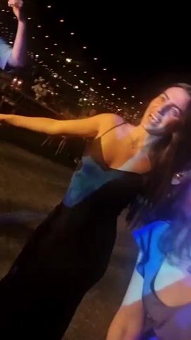 A low-angle, shaky video shows two women at night with blurred lights in the background. The woman in the foreground, with brown hair and wearing a purple shirt, is laughing with her mouth wide open. She is looking directly at the camera. Behind her, to her left, is another woman with long brown hair, wearing a black dress with a blue strap. She is also laughing and looking at the camera. The video is lit with blue and purple lights. The scene appears to be outdoors, possibly at a party or event.
