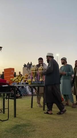 The video shows an outdoor award ceremony with a group of people gathered on a grassy field. In the foreground, a man wearing a dark vest over a traditional shalwar kameez and a white cap faces away from the camera, holding what appears to be a trophy. To his right, another man in a light blue shalwar kameez and white cap holds a piece of paper and looks at the man with the trophy. Behind them, a table displays several gold trophies. Several spectators are seated and standing in the background, some looking towards the ceremony. The sky is a pale orange, suggesting it is either sunrise or sunset.