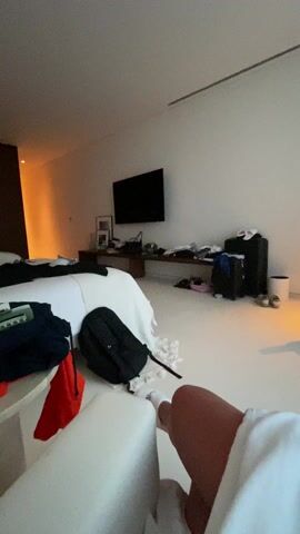The camera, held by a person sitting on a white chair, is looking around a messy hotel room. The room features a large bed with white sheets, a brown wooden accent wall, and a large TV mounted on a white wall. A small round table beside the chair holds a bottle of clear liquid, a water bottle, and several items of clothing. A black backpack is also on the table. Luggage can be seen on the floor, and various personal items are scattered throughout the room. The person holding the camera is wearing white socks and a white robe. Music is playing in the background.