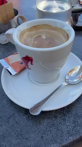 This media file features a close-up of a coffee cup filled with a creamy coffee, sitting on a saucer with a small biscuit packet and a spoon. The cup has a red dolphin logo and "Cremcaffè" written on it. Next to the coffee, there is a glass of water and a mobile phone. In the background, a person's legs and arm are visible, suggesting they are seated at a table.

The scene then pans out to reveal a beautiful coastal view. A low stone wall separates a paved walkway from the clear blue sea. In the distance, a hazy coastline with hills can be seen under a bright blue sky dotted with white clouds. The sunlight casts shadows on the walkway, indicating it is daytime, likely late morning or early afternoon, given the bright lighting. The overall impression is one of a relaxed, enjoyable moment, possibly a vacation or a leisurely outing by the sea. The presence of the coffee, water, and phone suggests a break or a moment of personal time. The location context indicates this is Malinska, Croatia. There are no discernible emotions expressed, but the setting evokes a sense of tranquility and pleasant weather.