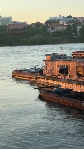 A slow-motion video shows a barge floating on a river. The barge is made of metal and appears to be old and rusted. It is covered with a tarp and has a small cabin on top. The river is wide and calm, and the water is a murky brown. In the background, there are trees and buildings on the shore. The sky is a pale yellow, indicating either sunrise or sunset. The sun's light casts a warm glow on the barge, highlighting its weathered texture.