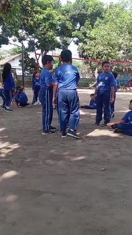 This is a medium-angle, slightly shaky, outdoor video showing a group of children, likely students, on a dirt court with scattered trees and a volleyball net in the background. Most of the children are wearing matching blue uniforms with white stripes down the sides of their pants and on the sleeves of their shirts. They appear to be engaged in some form of physical activity or game.

In the foreground, two boys in the blue uniforms are facing each other. One boy, on the left, has his hands behind his back and is looking at the other boy. The boy on the right is looking slightly away and has his right arm bent at the elbow.

To the left of these two boys, a girl in the same blue uniform is lying on her side on the ground, with her legs bent. Behind her, other children in uniform are partially visible.

In the middle ground, several other children in uniform are standing and looking around. One boy in the center, facing away from the camera, has a white logo or text on the back of his shirt.

To the right of the foreground boys, another boy in the blue uniform is standing and looking towards the left. Further back on the right, more children in uniform are visible, some sitting on the ground.

The ground is a dusty, sandy-brown dirt. Sunlight is filtering through the trees, creating dappled shadows. A white building is visible in the background behind some trees and the volleyball net. The overall atmosphere appears to be a casual, outdoor activity session.