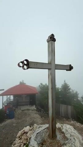 A group of people are visiting a mountain-top religious site in Dzirovani, Georgia, shrouded in fog. The scene features several crosses, with a prominent metal cross in the foreground. Behind it, a small wooden chapel with a red roof sits on a rocky outcrop. Two women and several other figures are visible, some standing near the chapel, others further back near a railing overlooking a misty valley. One woman is looking directly at the camera. The weather is overcast and foggy, creating a somewhat somber and mysterious atmosphere. The time of day appears to be daytime, but the fog obscures clear visibility and a distinct sense of light. The presence of the crosses and chapel indicates a spiritual or pilgrimage location. Blue barrels are visible near the chapel, possibly for storage or waste. A wooden fence can be seen on the right side of the image.